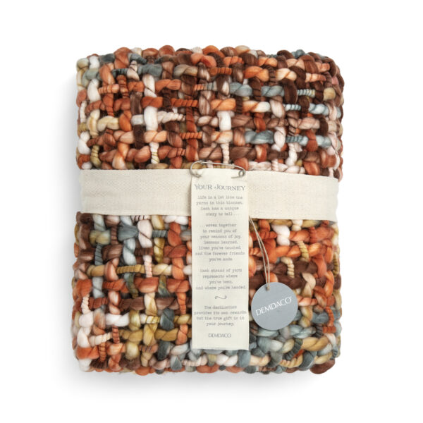 A woven fringed blanket with warm red, orange and cream colors, displayed folded and wrapped with cream ribbon and product tags.